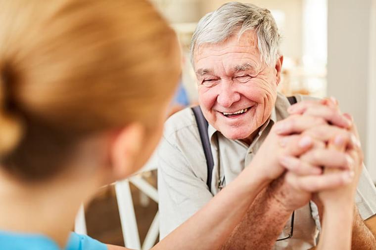 A carer holding the hands of a smiling older man in his living room